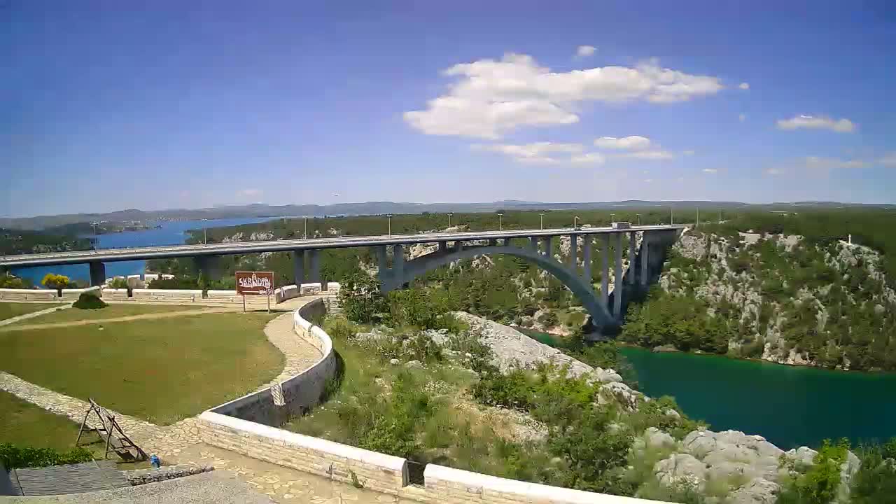 A long bridge spans a deep green river surrounded by lush green vegetation under a mostly sunny sky with a few fluffy clouds.