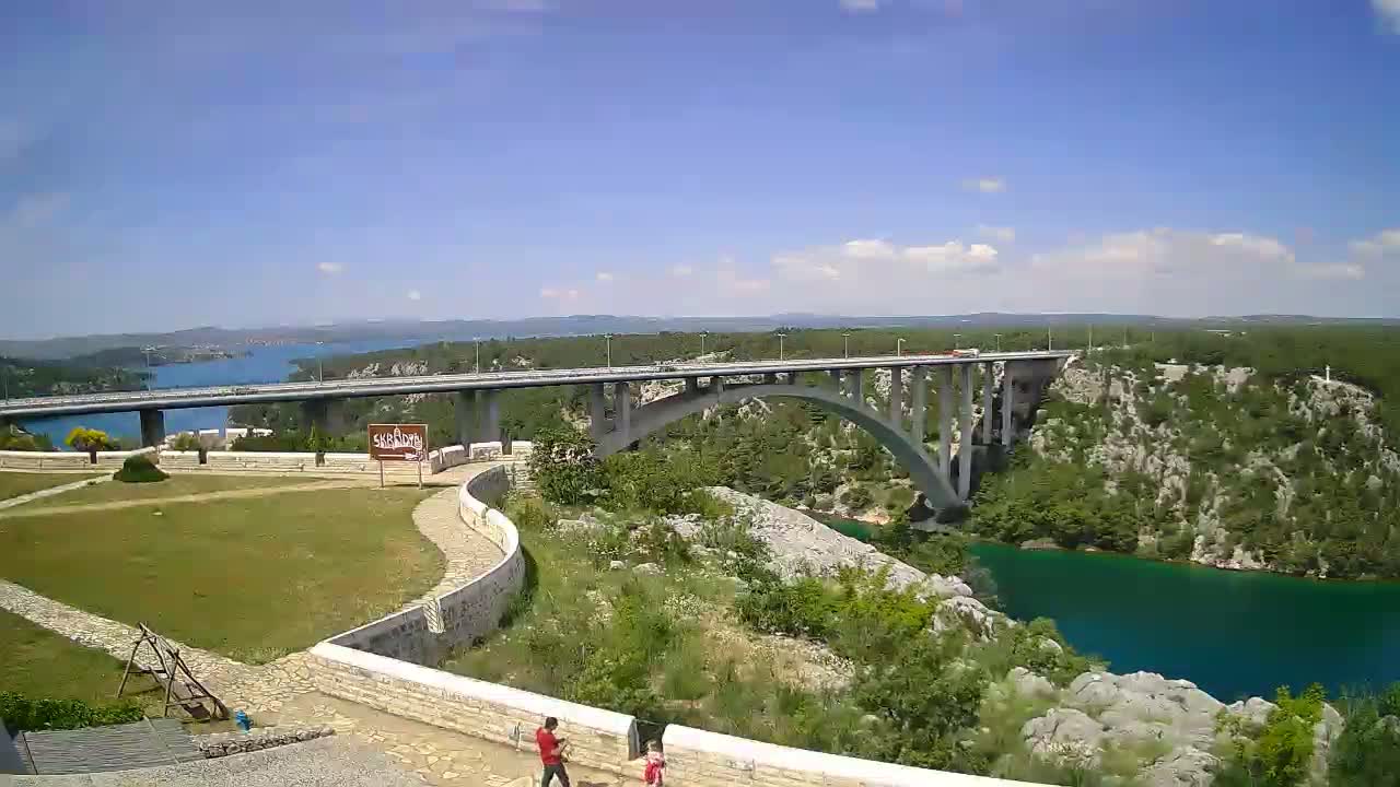 A long bridge spans a turquoise river, under a mostly sunny sky, with green vegetation lining the riverbanks and a grassy area in the foreground.