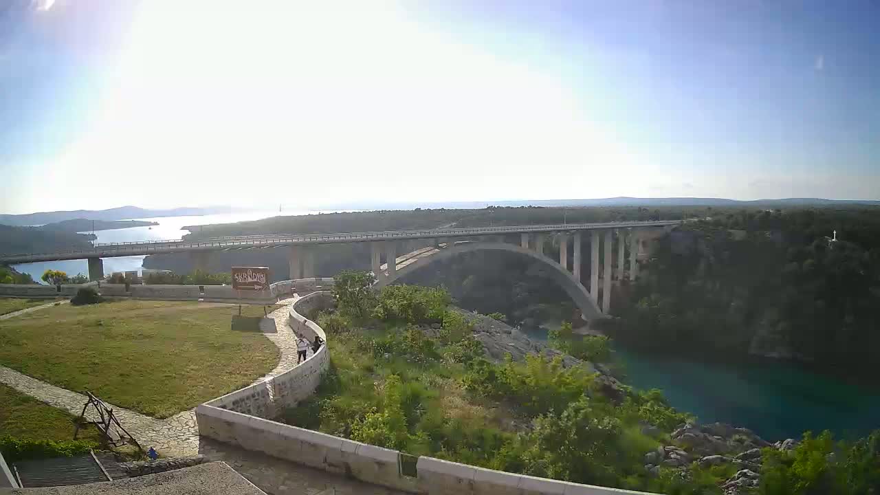 A long bridge spans a waterway on a sunny day, with green vegetation surrounding the water and a grassy area visible in the foreground.