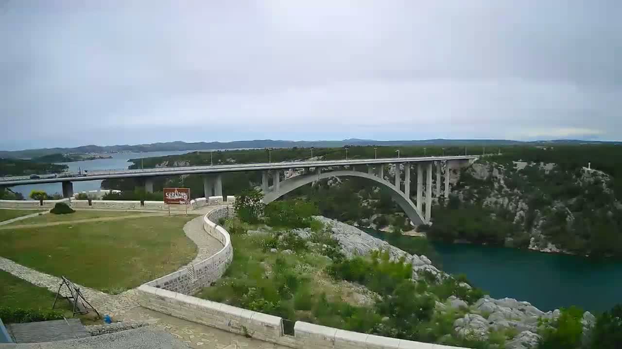 A long bridge arches over a teal river, surrounded by green vegetation under a cloudy sky.