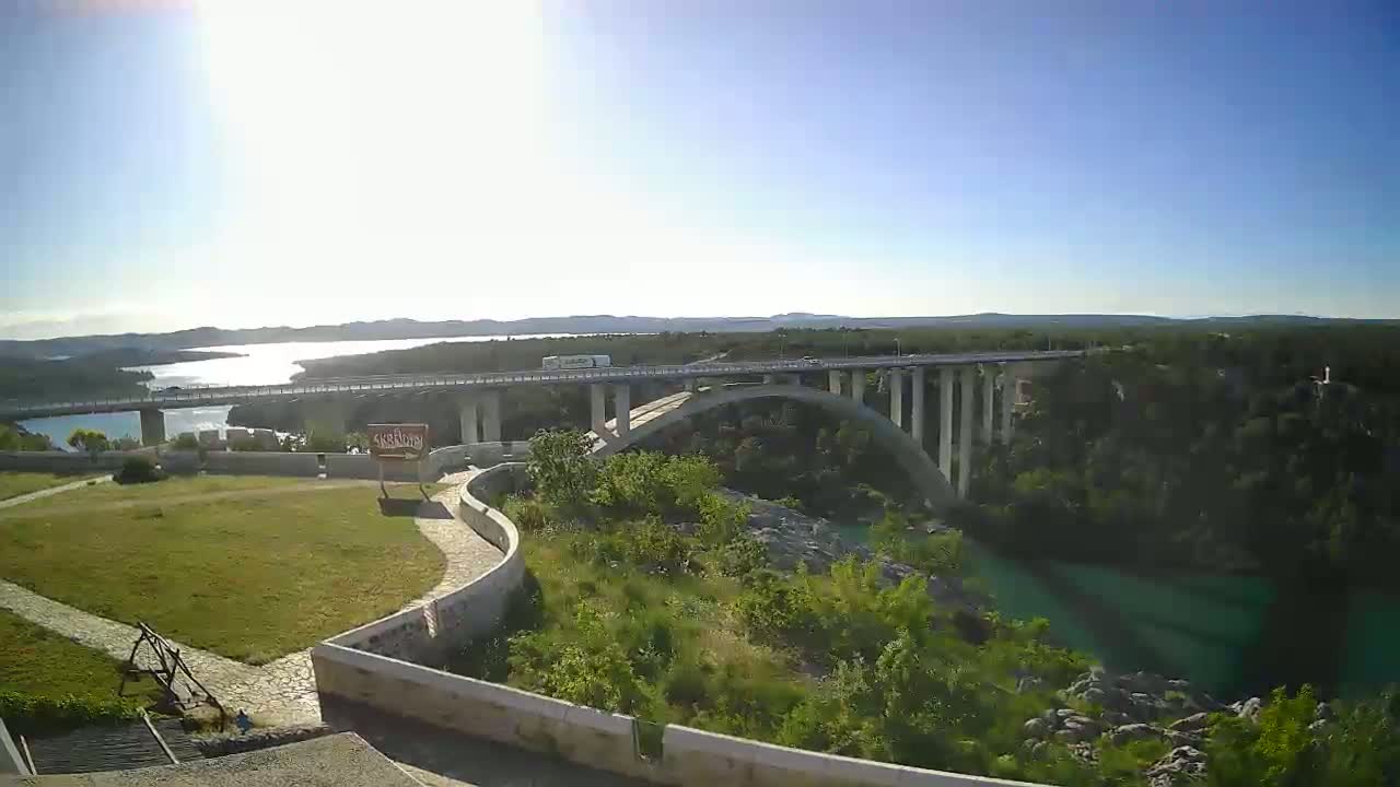 A long bridge arches over a teal river, with a sunny, clear sky overhead, and green vegetation surrounding the river and bridge.