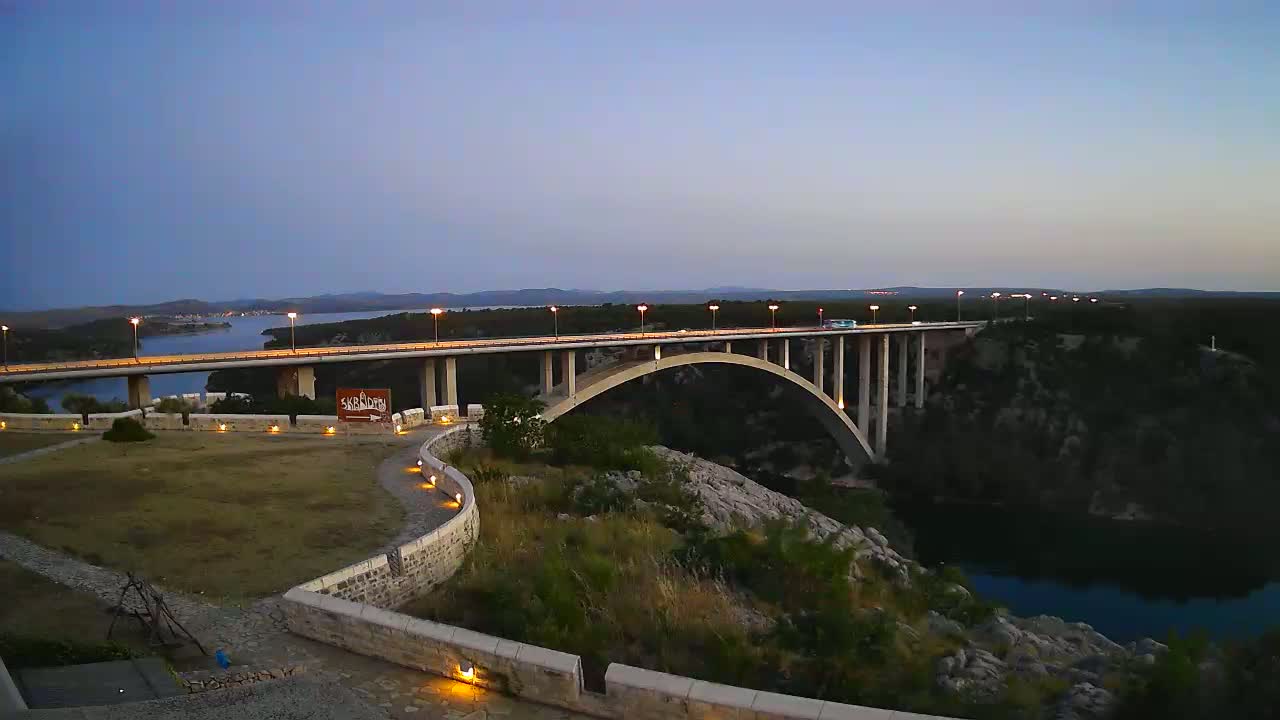 A long bridge with cars driving across it at dusk, spanning a dark body of water surrounded by hilly, dark green and rocky terrain.