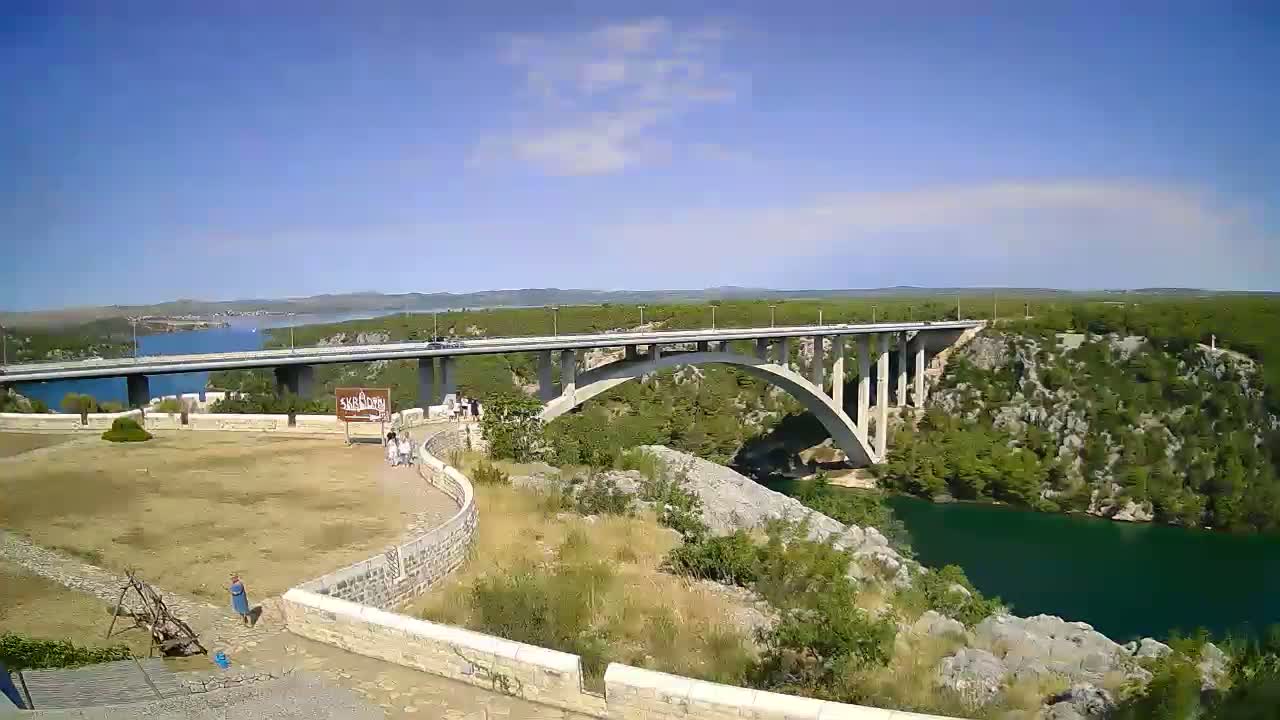 A long concrete bridge spans a wide, green river under a clear blue sky with a few scattered clouds.