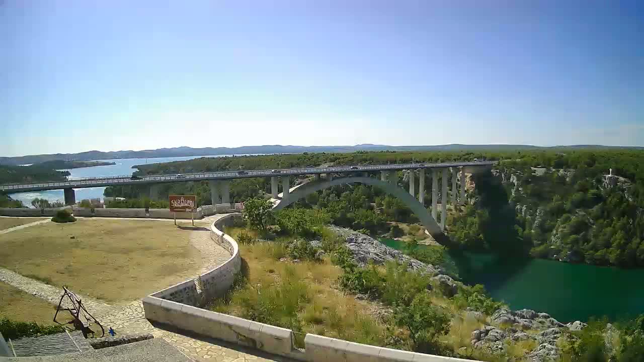 A long concrete bridge spans a wide, green river under a clear blue sky with a few scattered clouds.