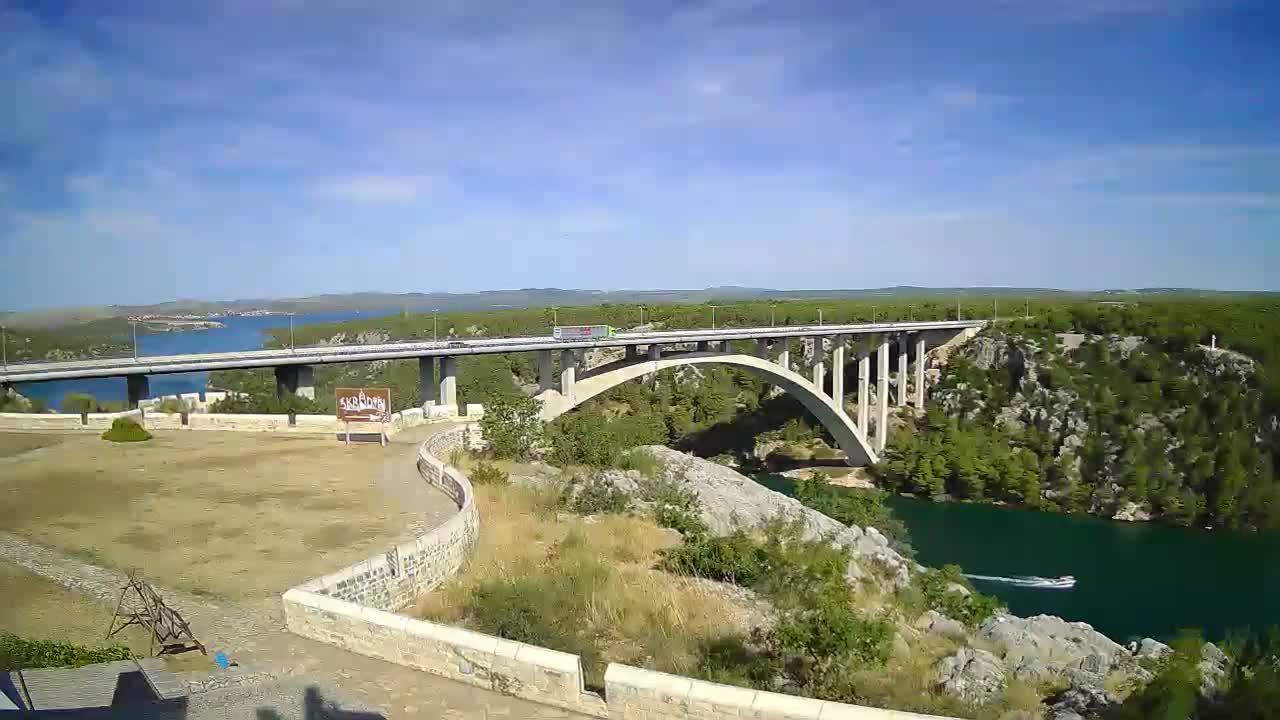 A long bridge spans a river, with a small boat visible on the water under a mostly sunny sky.