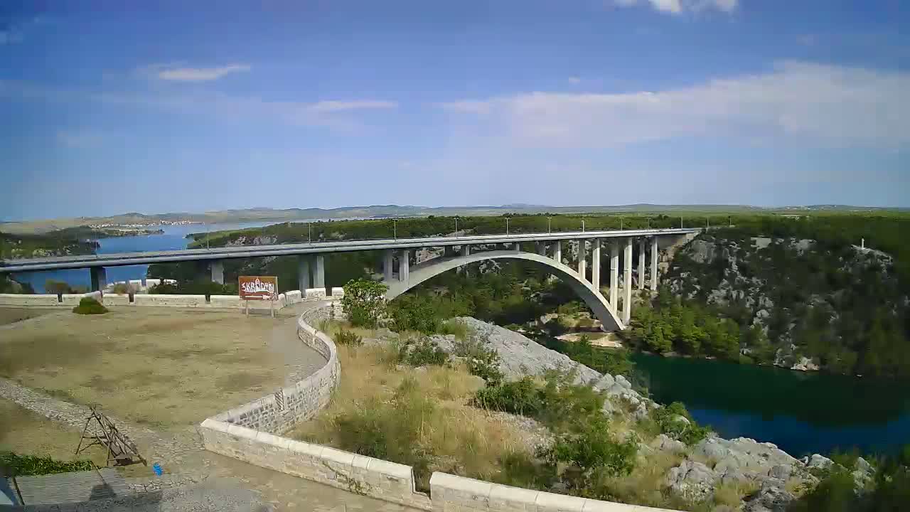 A long concrete bridge arches over a blue river, under a mostly sunny sky with some scattered clouds.