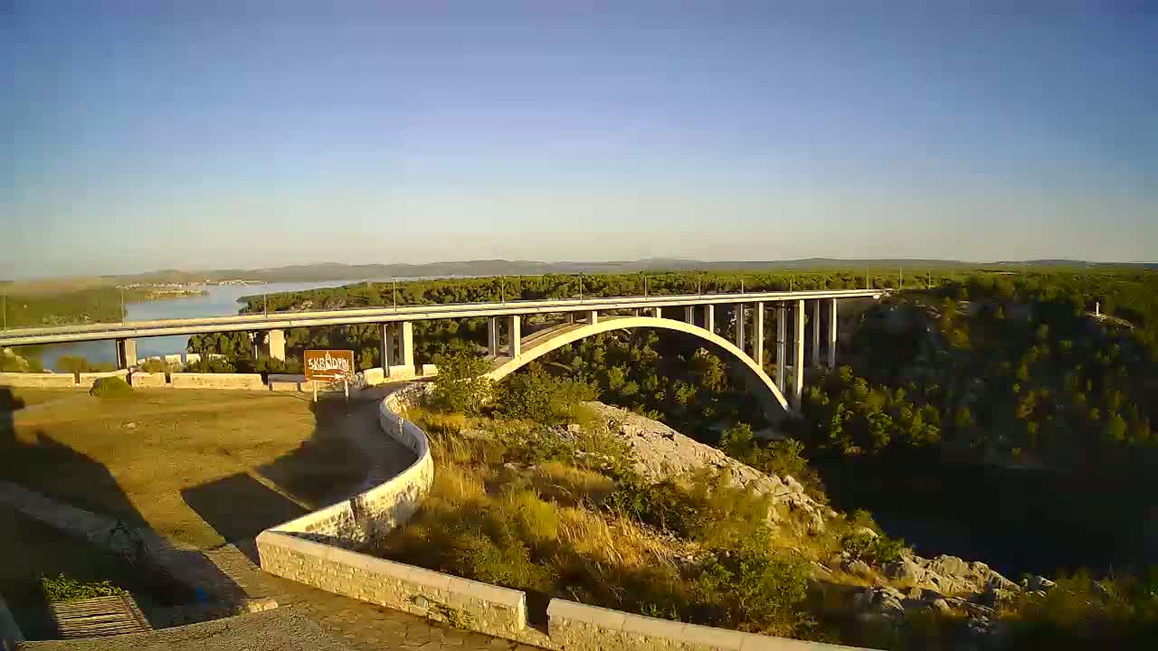 A long concrete bridge arches over a blue river, under a mostly sunny sky with some scattered clouds.