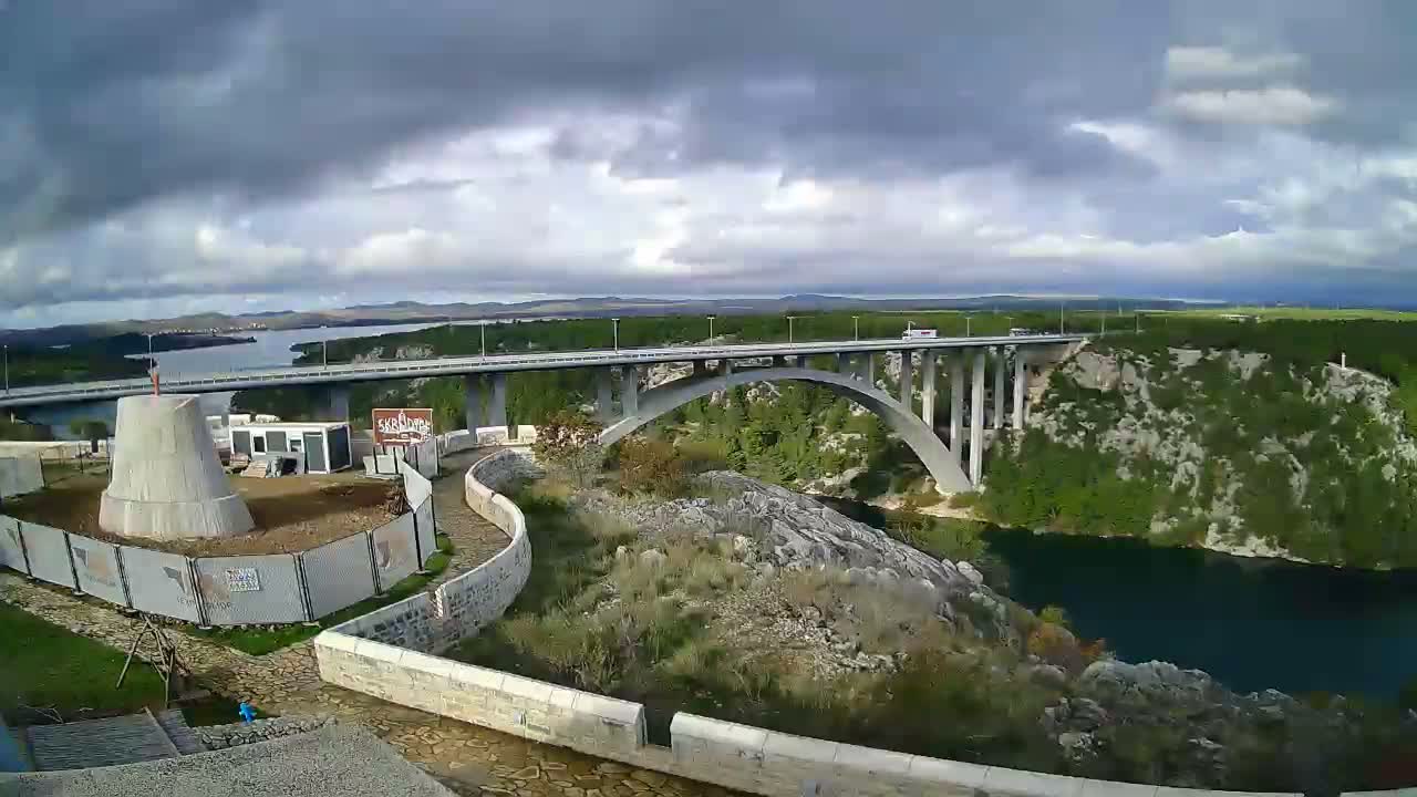 A high-angle view reveals a multi-arched concrete bridge crossing a deep river gorge amidst rocky, green hills, with a conical structure under construction in the foreground, under a partly cloudy sky with dramatic grey clouds.