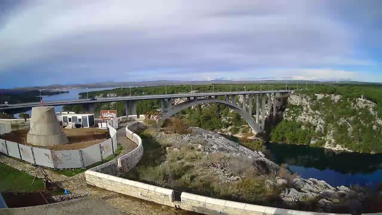 A wide view captures a large arched bridge spanning a deep river canyon surrounded by forested hills and rocky terrain under a partly cloudy sky, with a construction site visible in the foreground.