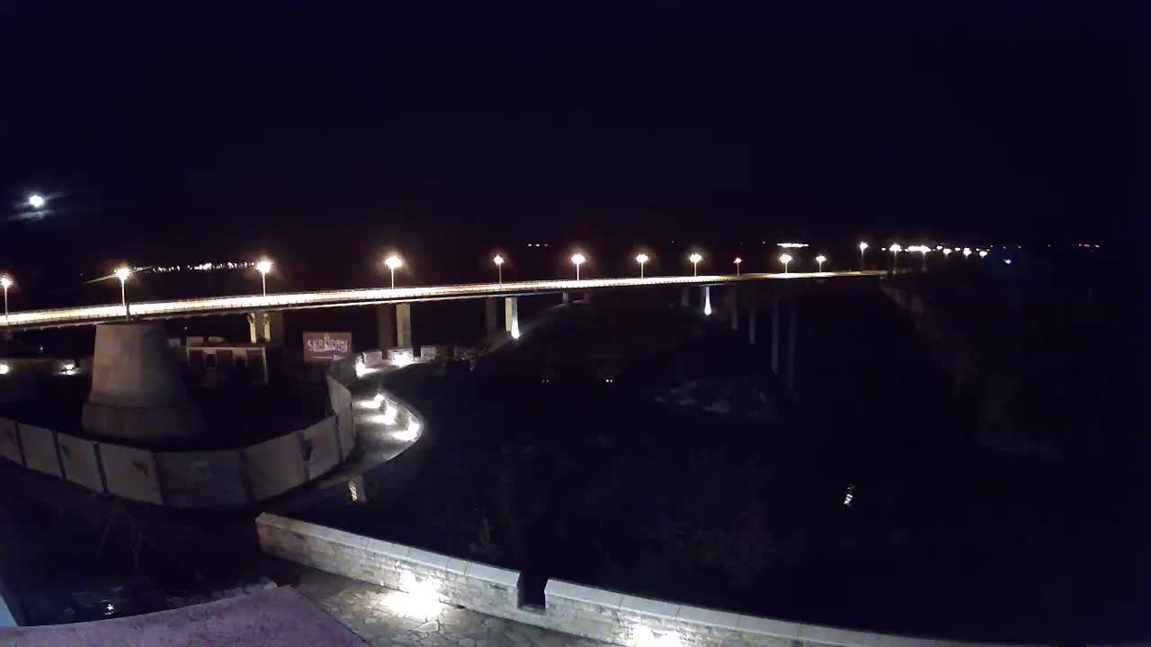 A long, illuminated bridge stretches over dark water under a clear, moonlit night sky, with a curving lit path in the foreground.