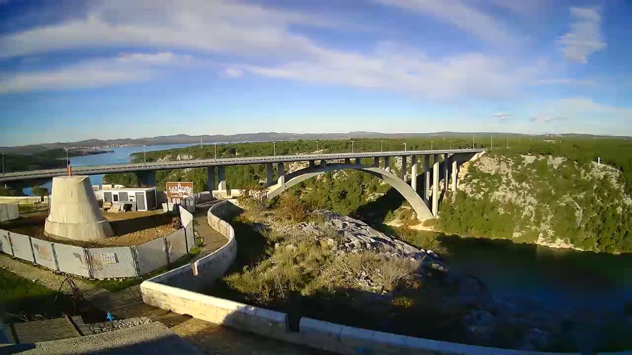 A wide-angle outdoor shot captures a long concrete arch bridge crossing a deep green canyon or river leading to a wider body of water, framed by rocky hills covered in green vegetation, with a conical concrete structure in the foreground, all under a bright blue sky with scattered white clouds.