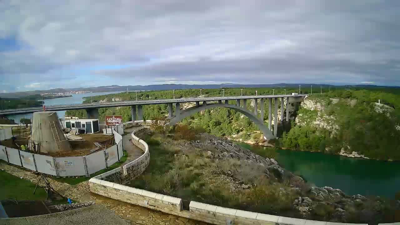 A large concrete arch bridge spans a greenish river surrounded by tree-covered hills and a distant town, all under a mostly cloudy sky, with a construction site and winding path visible in the foreground.