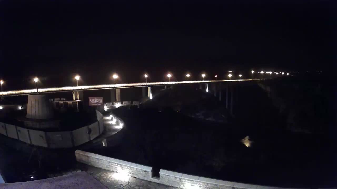 A long, well-lit bridge stretches over a dark expanse, with a curved illuminated pathway and wall in the foreground, all under a clear night sky.