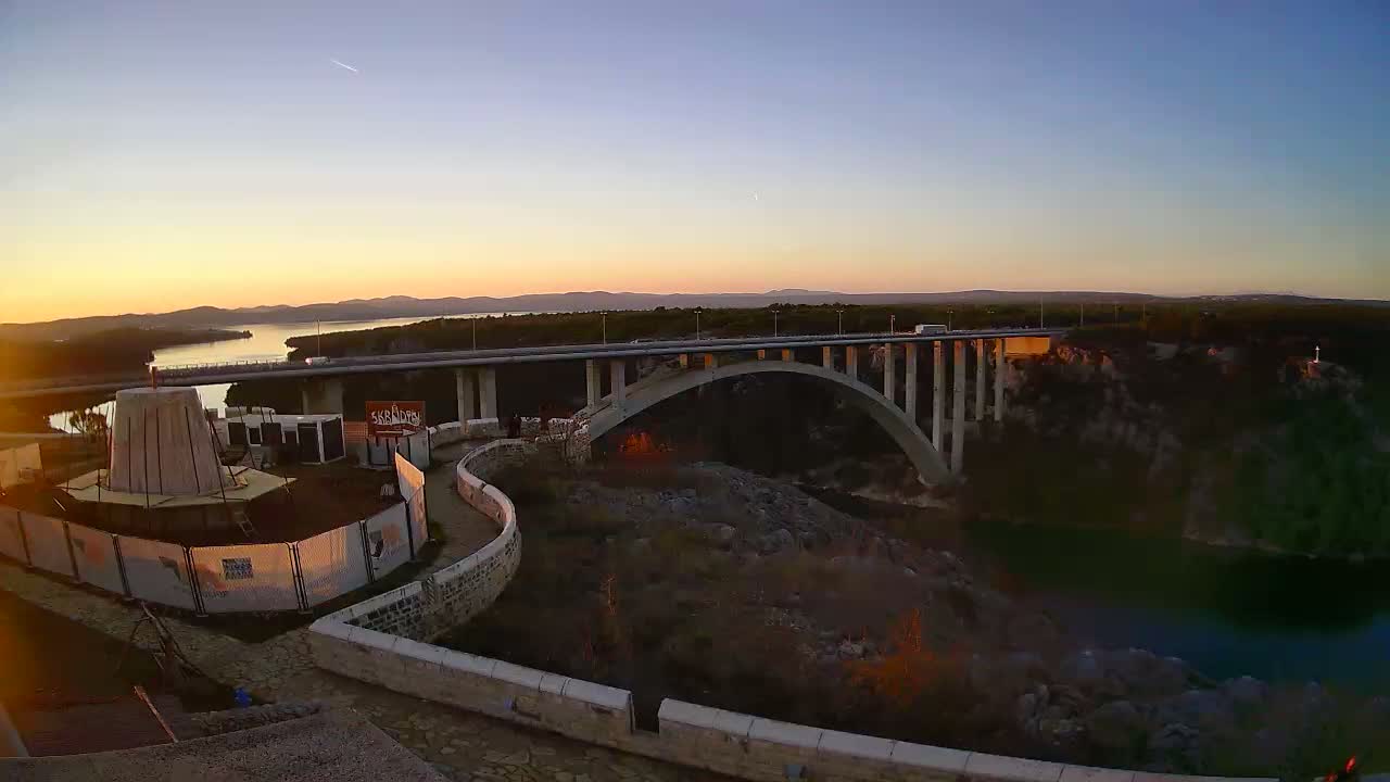 A long, well-lit bridge stretches over a dark expanse, with a curved illuminated pathway and wall in the foreground, all under a clear night sky.