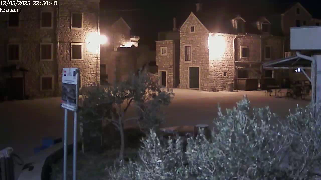 At night, stone buildings are illuminated in a square, surrounded by low shrubs.