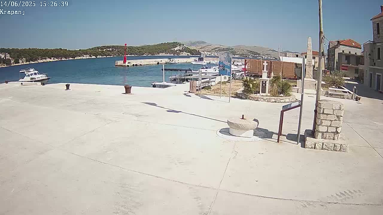 A sunny harbor scene shows a paved plaza next to a calm body of water with several boats and a small town in the background.