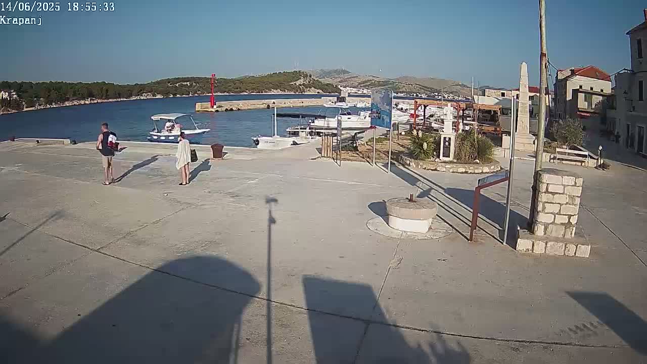 A sunny harbor scene shows a paved plaza next to a calm body of water with several boats and a small town in the background.