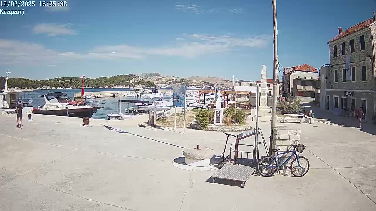 A sunny harbor scene shows a paved plaza next to a calm body of water with several boats and a small town in the background.