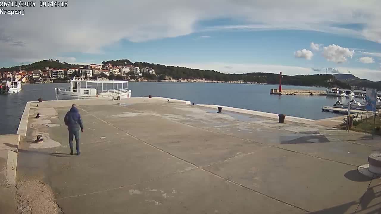 A person walks on a sunny concrete pier in a calm bay, with boats, a coastal town, and green hills visible under a partly cloudy blue sky, and a red lighthouse marking a small marina to the right.