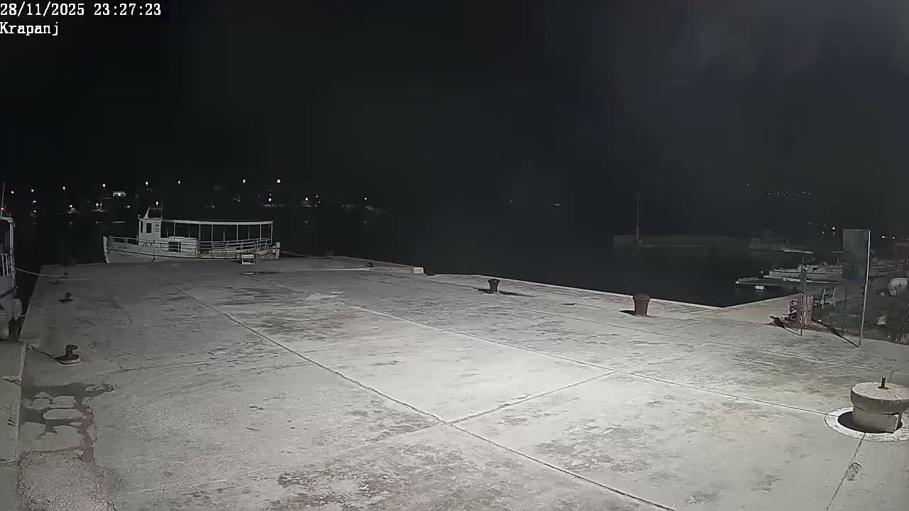 A concrete pier extends into a calm harbor at night, with several boats moored along the waterfront and distant lights visible across the water under clear conditions.