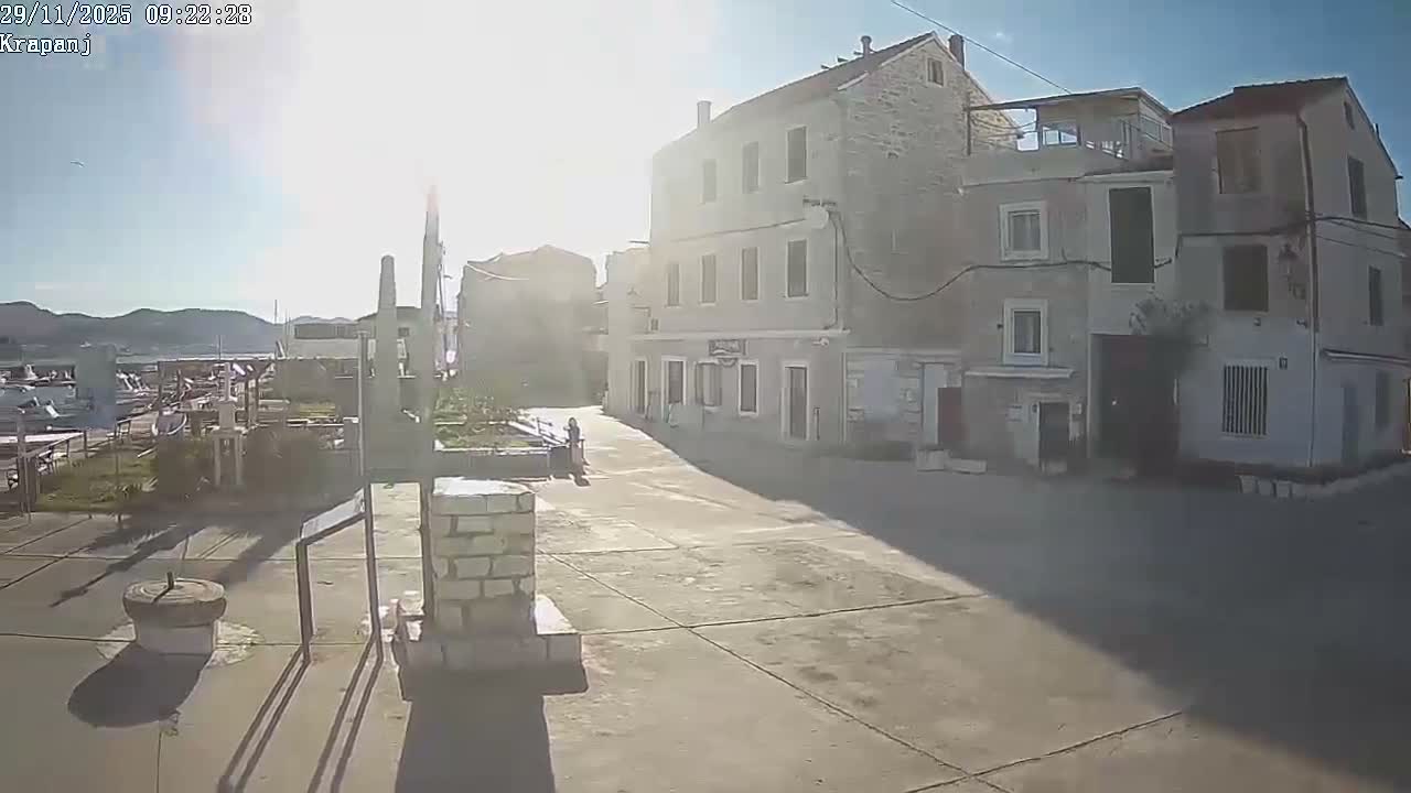 A bright, sunny day illuminates a paved town square featuring traditional stone buildings, a lone person, and a harbor filled with boats against a mountainous background.