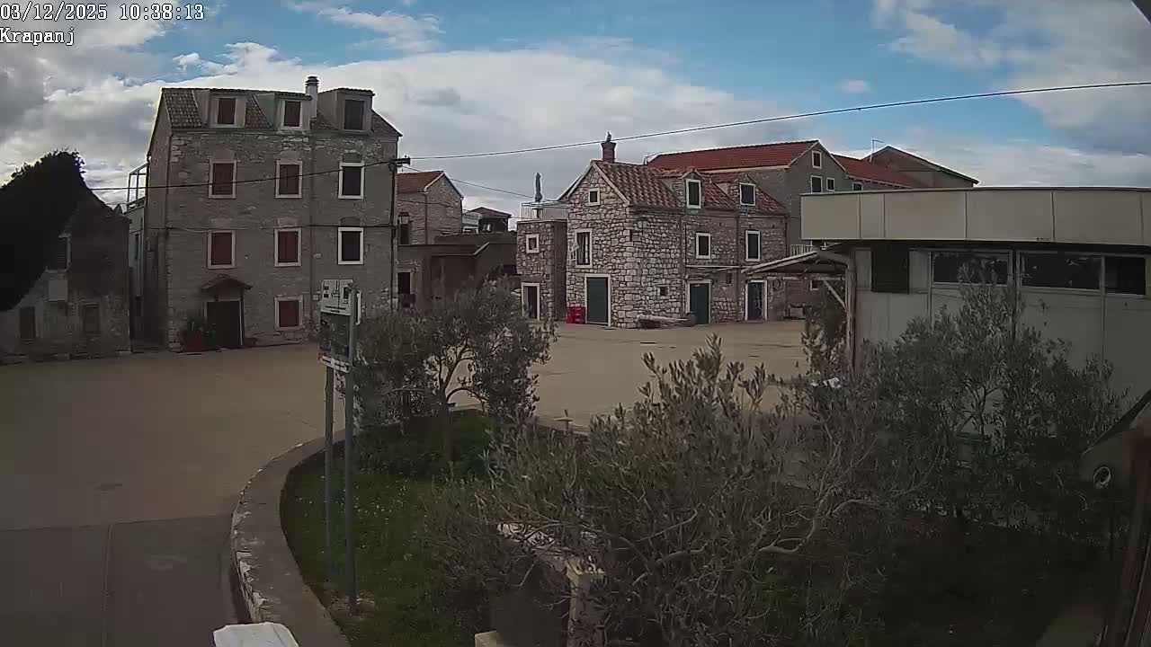 A quiet village scene features traditional stone buildings and a paved square with scattered olive trees under a partly cloudy sky.