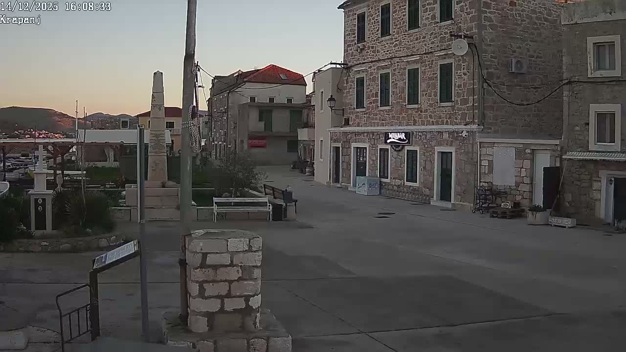 A quiet village scene features traditional stone buildings and a paved square with scattered olive trees under a partly cloudy sky.
