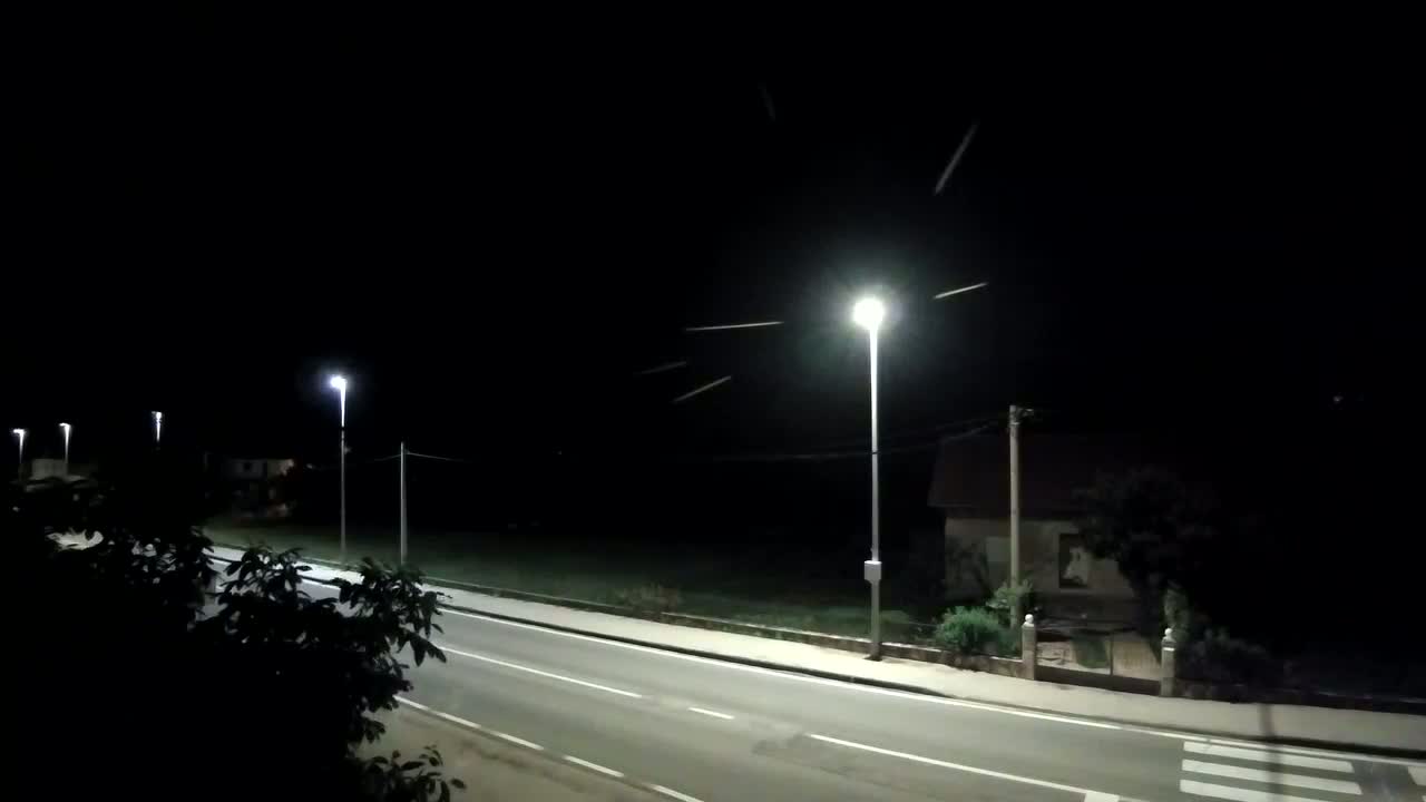 Under a night sky streaked with multiple meteors, a dark road illuminated by streetlights curves past a house and some vegetation.
