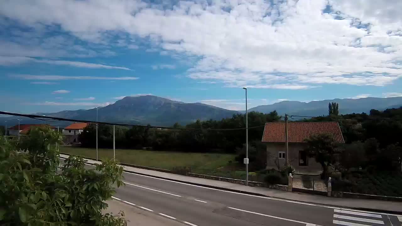 A paved road with a pedestrian crossing runs through a rural area beneath a partly cloudy blue sky, with mountains in the background.
