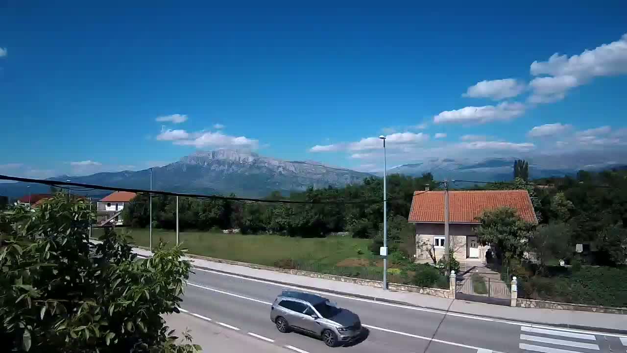 A silver SUV drives on a road past a house with a red roof, under a mostly sunny sky with some clouds, and mountains in the background.