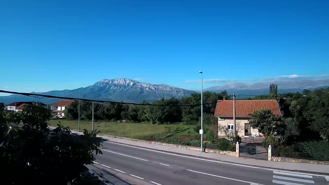 A paved road leads toward a small house with a terracotta roof, situated before a line of trees and a mountainous backdrop under a clear blue sky.