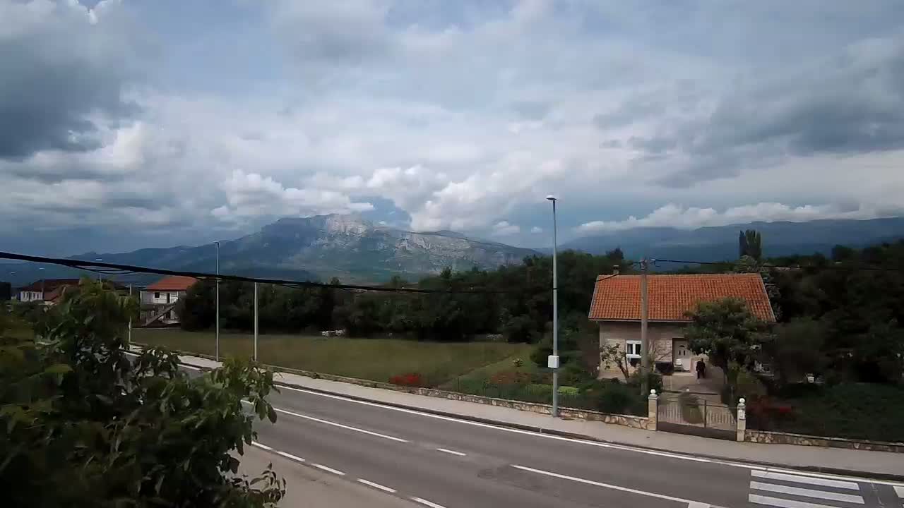 A paved road runs past a house with a red roof, situated amidst greenery and trees, with mountains under a cloudy sky in the background.