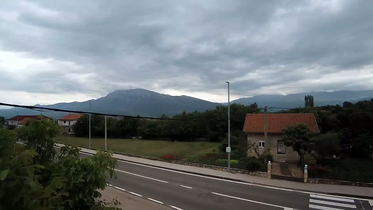 A two-lane road runs past houses and green space, with mountains in the background under a cloudy sky.