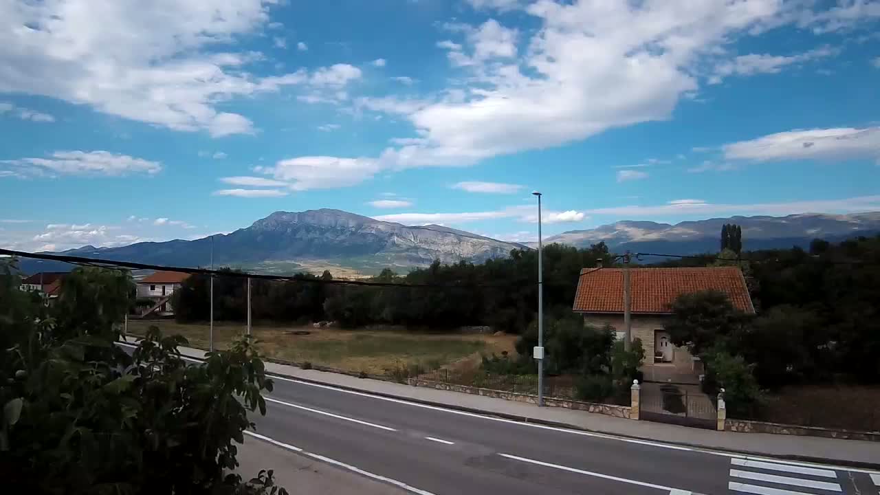 A two-lane road runs past houses and green space, with mountains in the background under a cloudy sky.