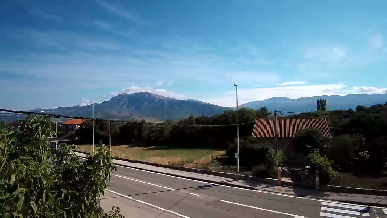 A paved road stretches toward a mountain range under a mostly clear, sunny sky, with a few scattered clouds and vegetation flanking the road.
