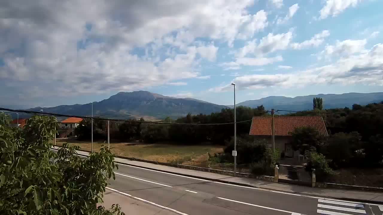 A partly cloudy sky overlooks a road and houses nestled amongst green trees and a mountain range in the distance.