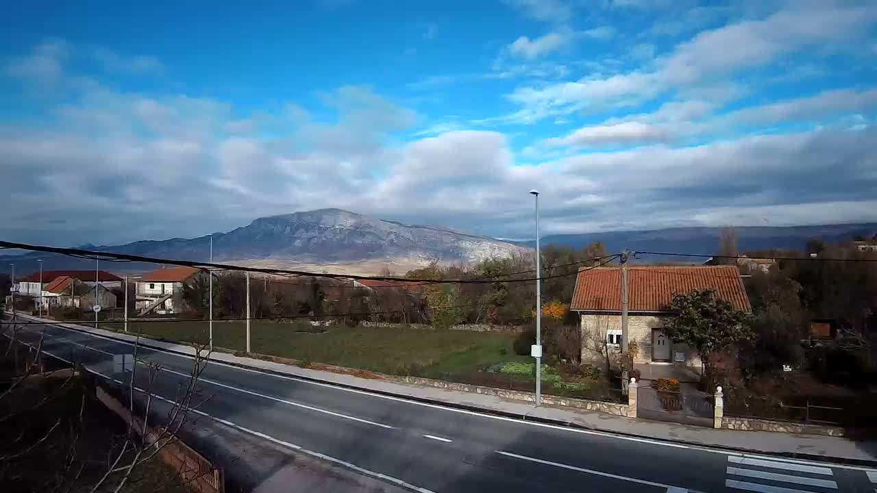 A partly cloudy sky overlooks a road and houses nestled amongst green trees and a mountain range in the distance.