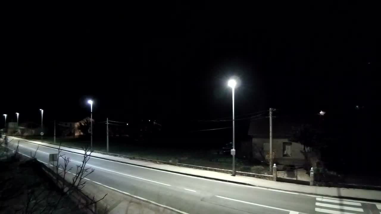 A partly cloudy sky overlooks a road and houses nestled amongst green trees and a mountain range in the distance.