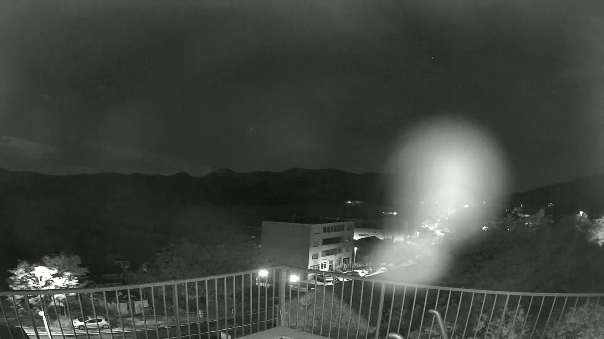 A nighttime view from a balcony shows a building and a parking lot below, with mountains in the distance under a dark, clear sky.