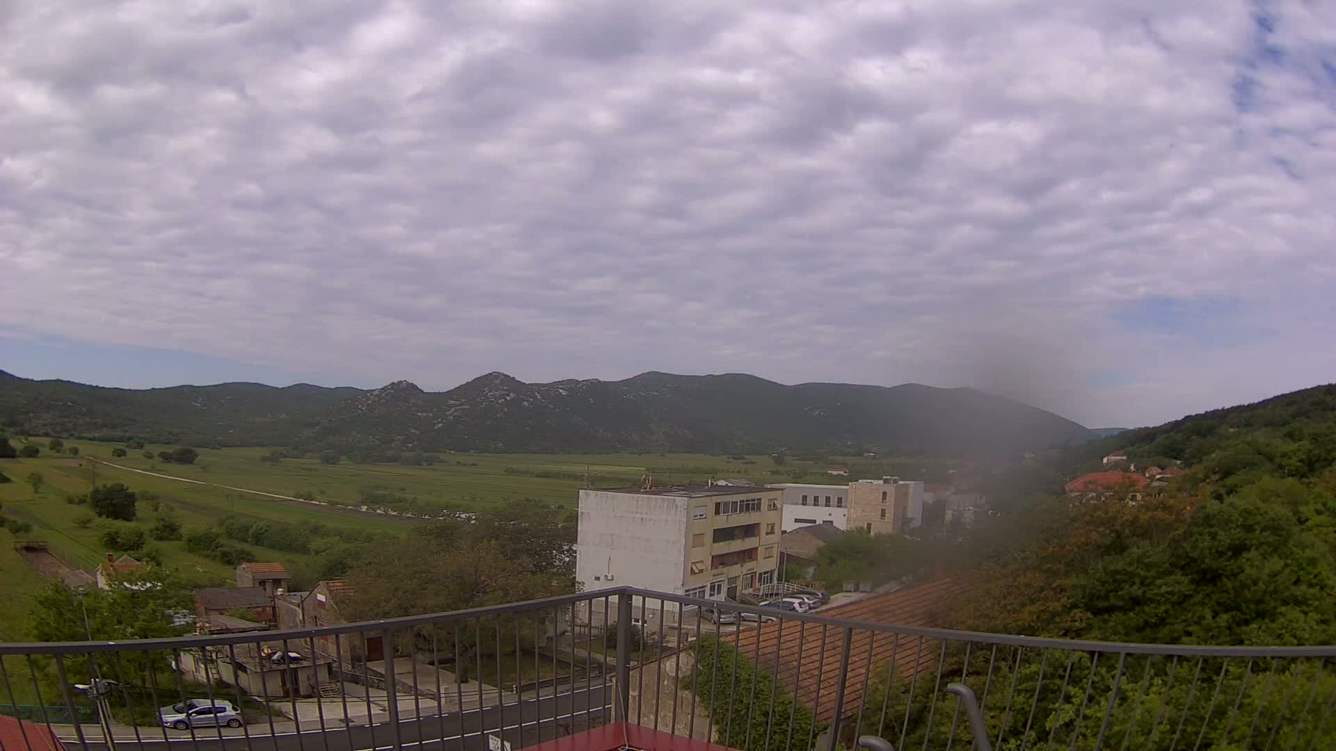 A view from a balcony shows a town nestled in a valley between green hills under a cloudy sky.