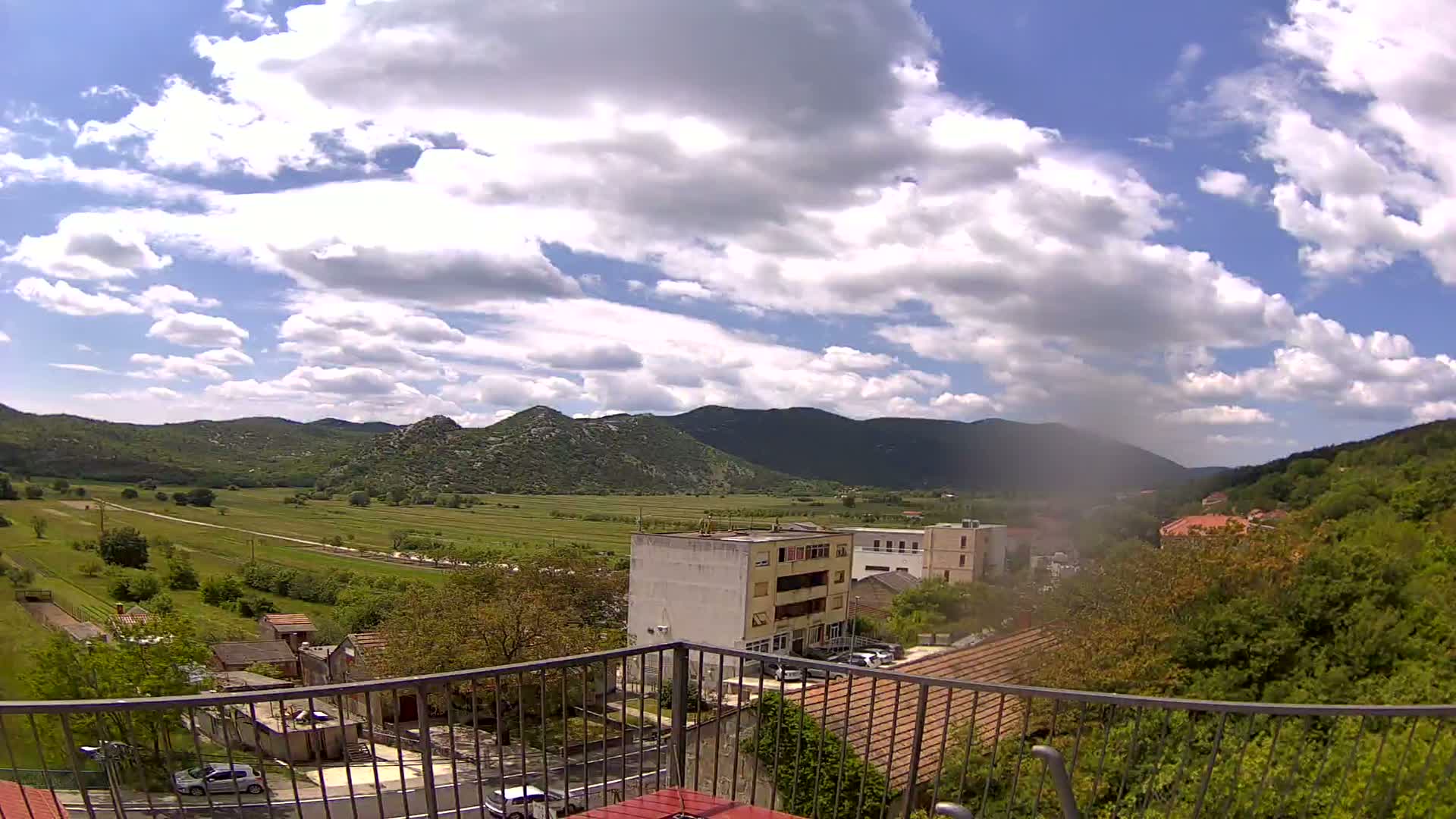 A partly cloudy day reveals a view of a valley with buildings, green fields, and mountains in the background, seen from behind a metal balcony railing.