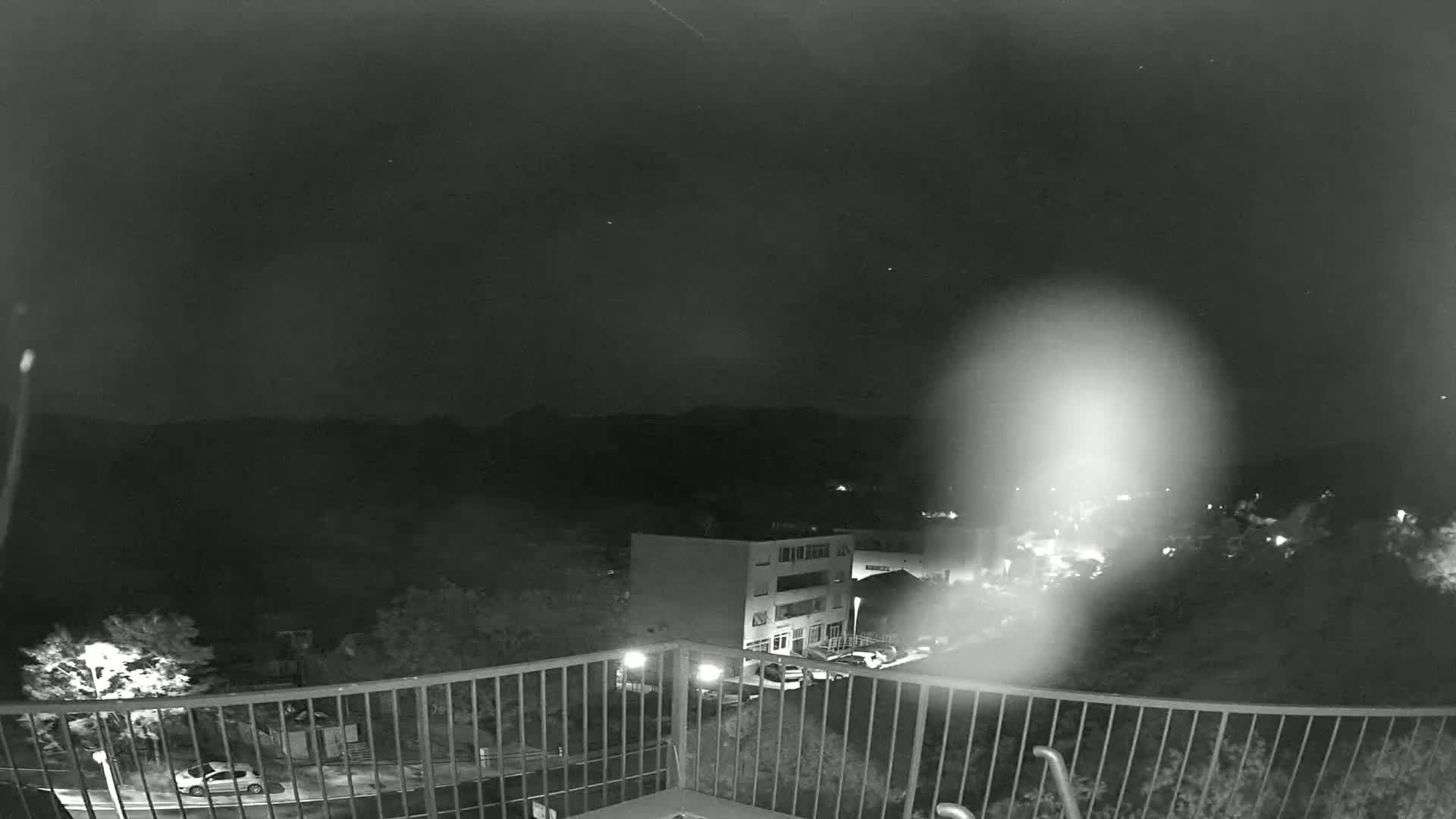 A nighttime, low-light view from a balcony shows a building and parked cars below, with hills in the distance under a clear, starry sky.