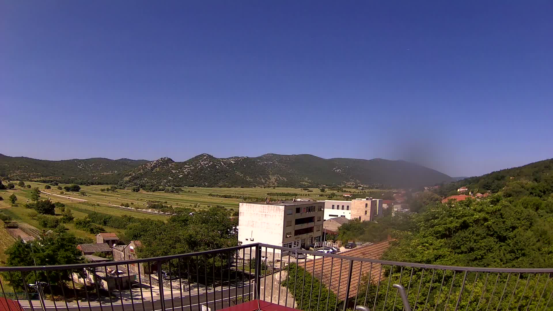 A sunny day reveals a valley with farmland, buildings, and low, tree-covered mountains in the distance, viewed from behind a balcony railing.