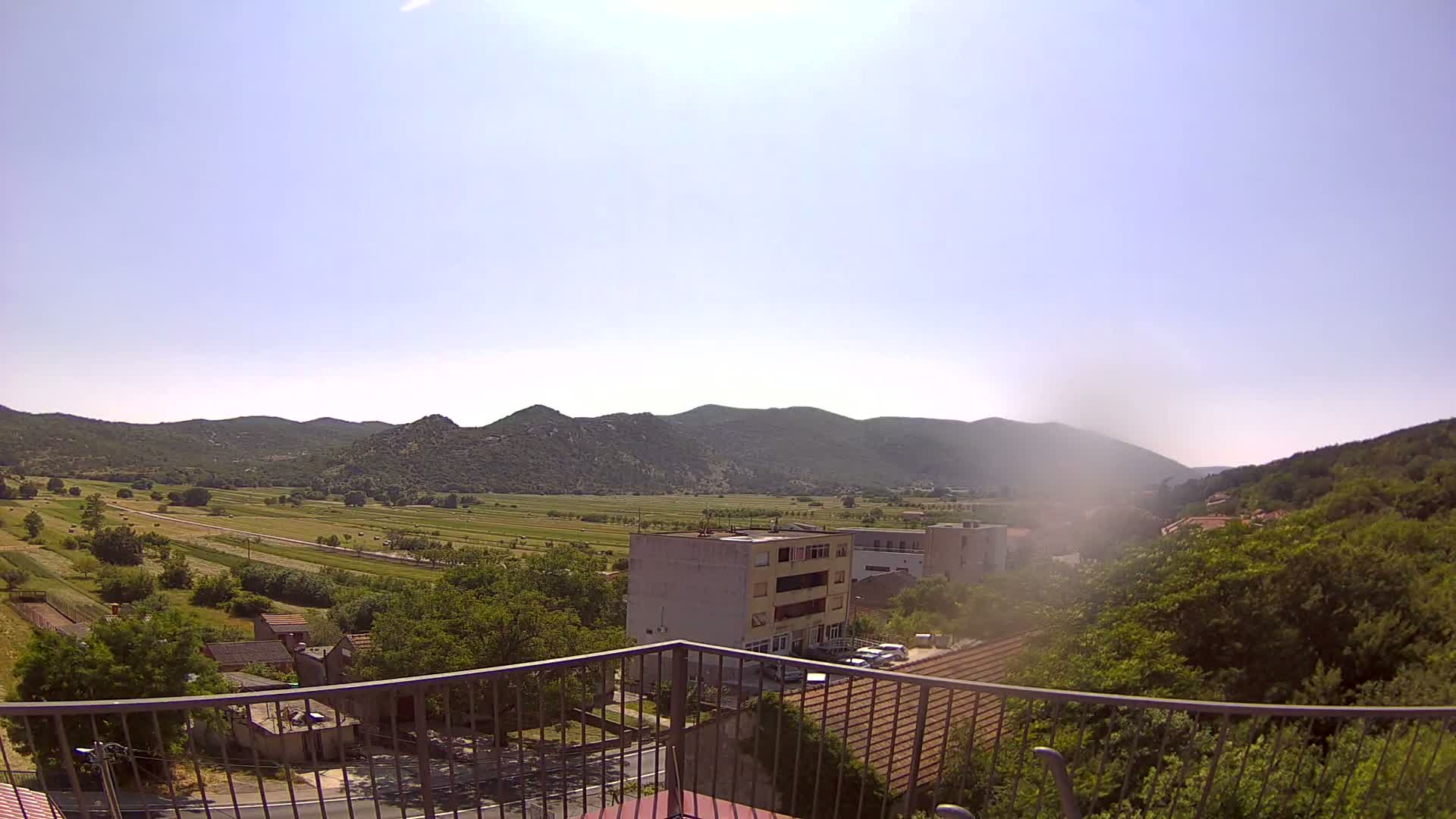 A sunny view from a balcony shows a valley of green fields and houses, nestled between low, rocky mountains.