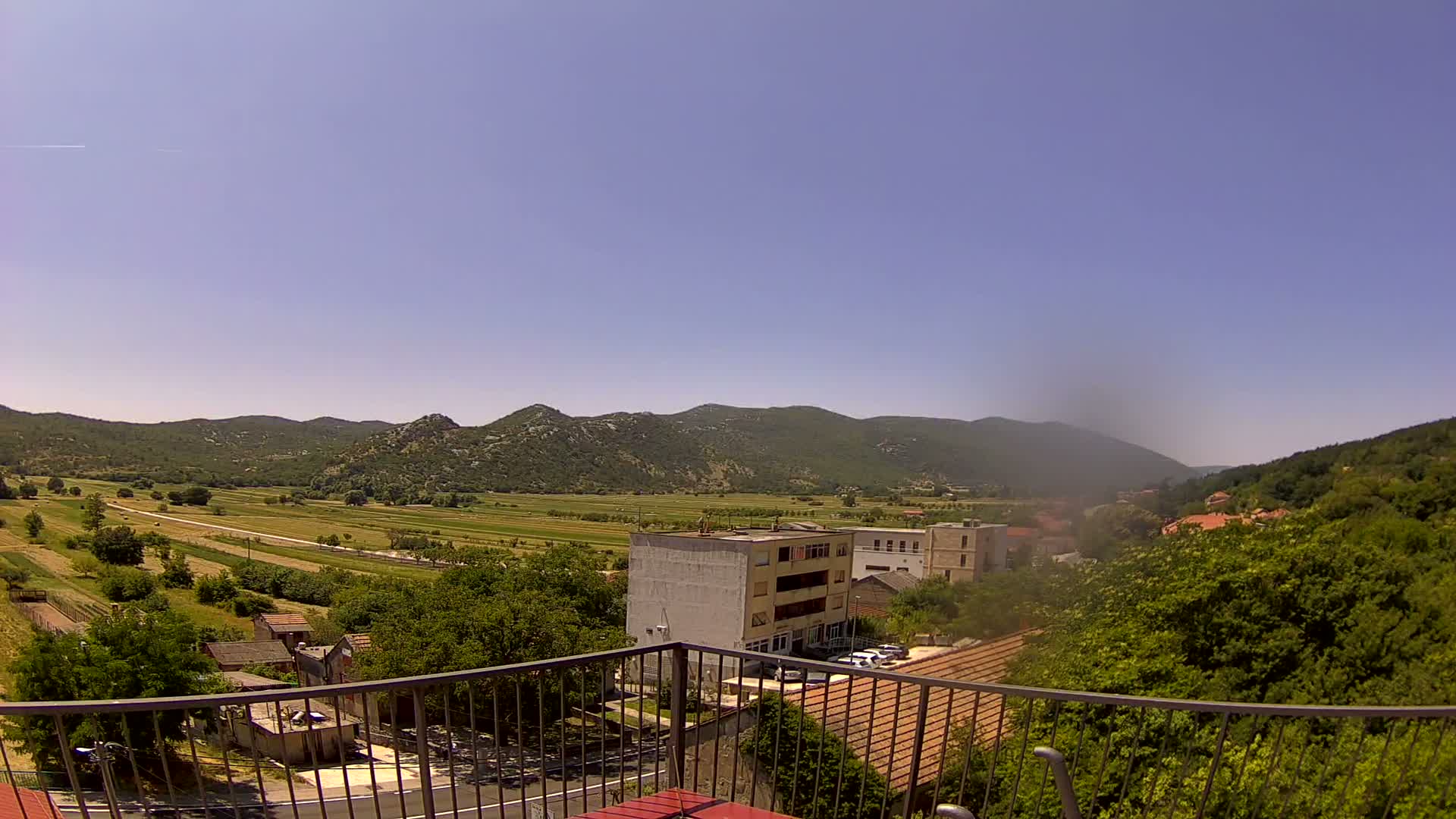 A sunny view from a balcony shows a valley of green fields and trees, bordered by low, rounded mountains, with a few buildings visible in the mid-ground.