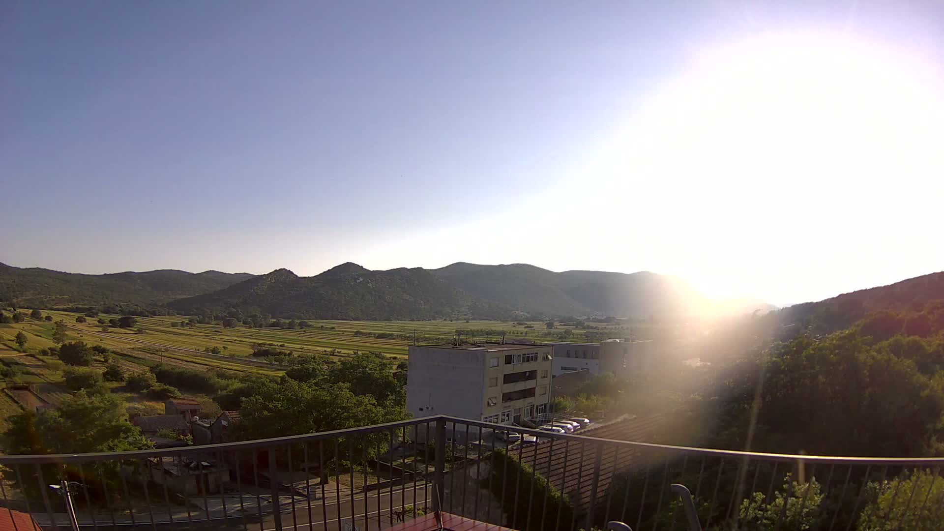 A sunny view from a balcony shows a valley of green fields and low mountains under a clear blue sky.