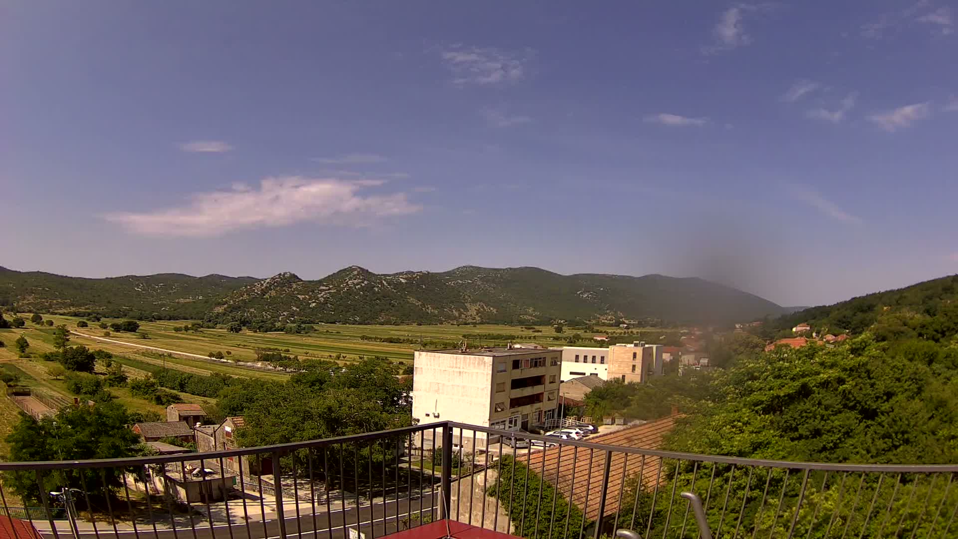 A mostly sunny view from a balcony shows a valley of farmland and a small town nestled against low, green mountains.