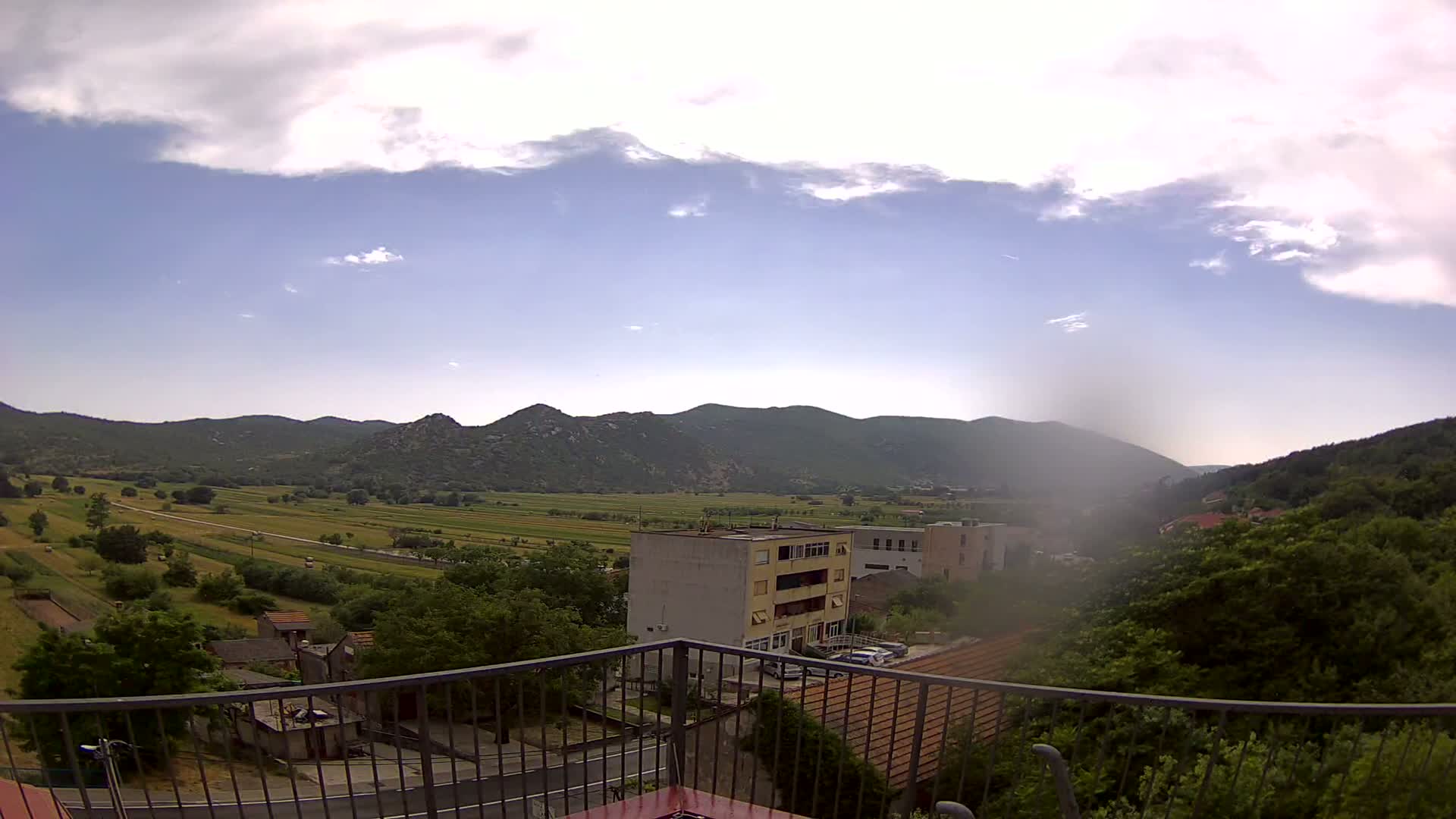 A partly cloudy day reveals a valley view of green fields, buildings, and mountains from a balcony with a metal railing.