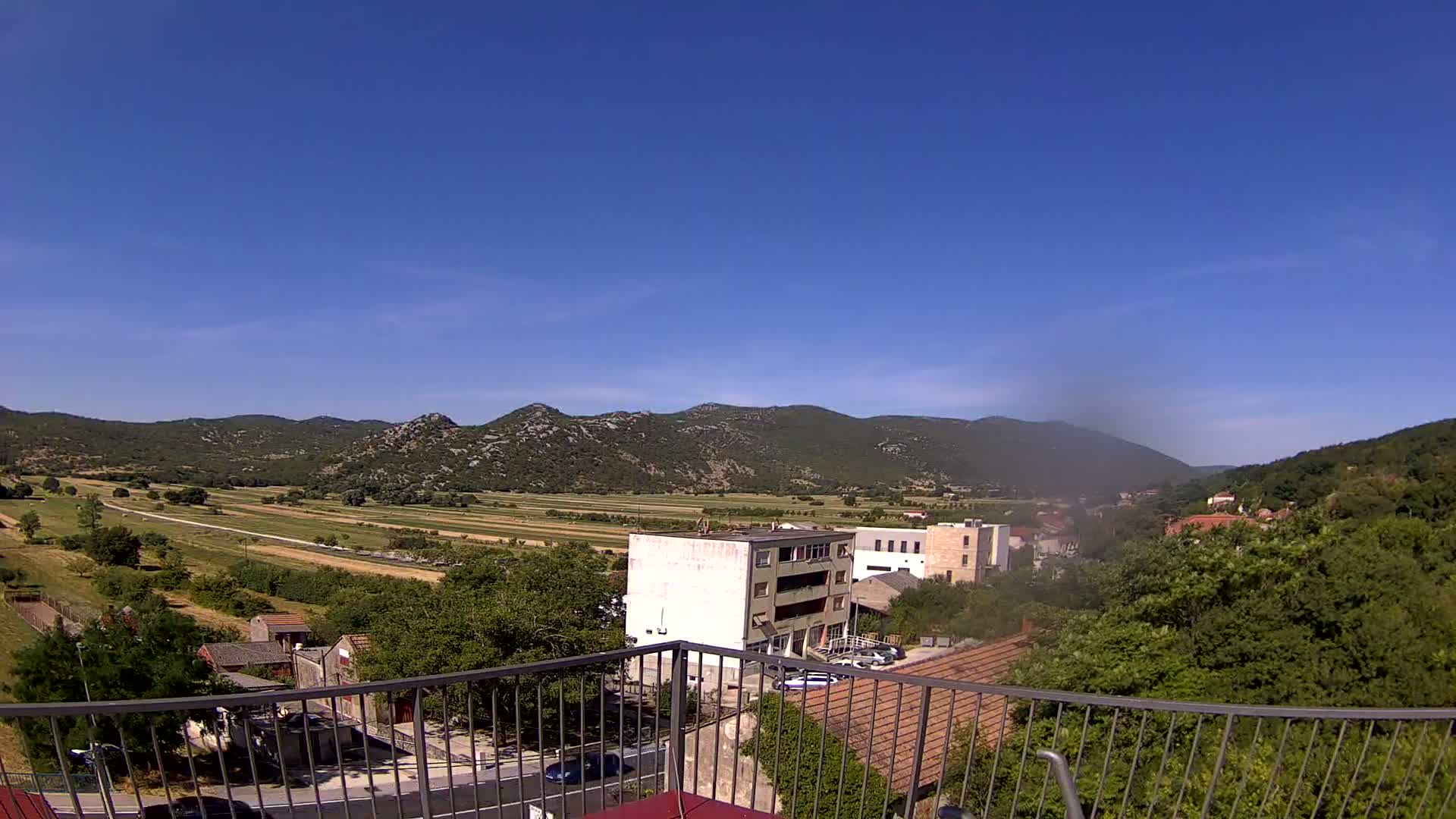 A sunny day view from a balcony shows a valley of green fields and low buildings nestled between hills.