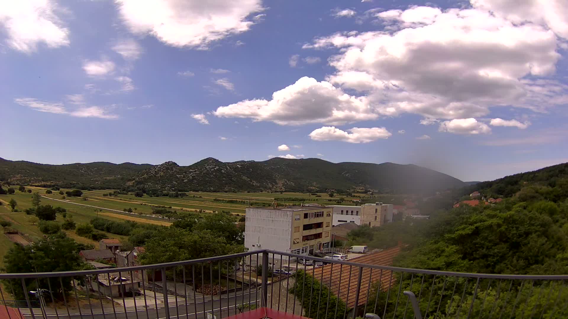 A sunny day view from a balcony shows a valley of green fields and low buildings nestled between hills.